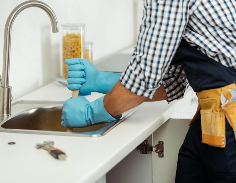 plumber-in-tool-belt-and-rubber-gloves-cleaning-blockage-of-kitchen-sink